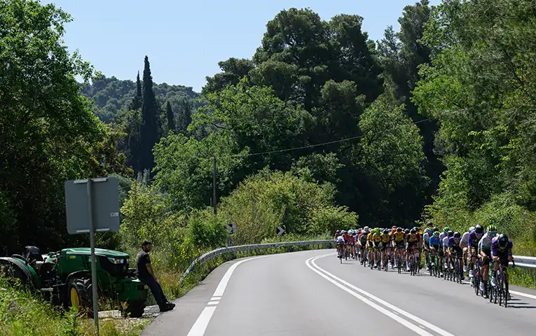 A farmer is sitting on a tractor on the side of the road while cyclists are cycling through the trees during DEI International Tour of Hellas